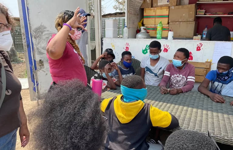 A group of people, including children wearing masks, sit around a table outside while an adult in a red shirt and cap stands nearby, giving instructions.