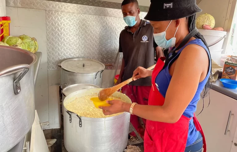 Two individuals wearing masks prepare food in a kitchen. One person in a red apron and hat is ladling food from a large pot into a bowl, while the other stands nearby in the background.