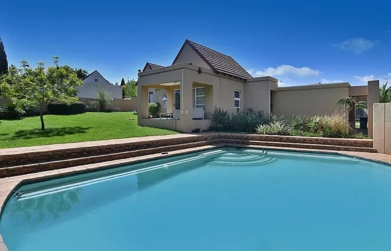 A swimming pool with clear blue water in a backyard, adjacent to a small house with a gable roof and covered patio. Lush green lawn and a tree are visible in the background.