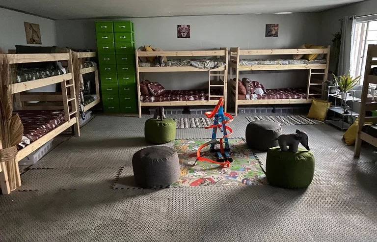 A neatly organized room with four wooden bunk beds, green lockers, a play mat with a spiral toy, and several round cushions. The room has gray flooring and natural light from the window to the right.