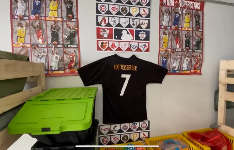 A football jersey with the name "Roethlisberger" and the number 7 is displayed on a wall with various sports posters and team logos above a green storage bin and yellow container.