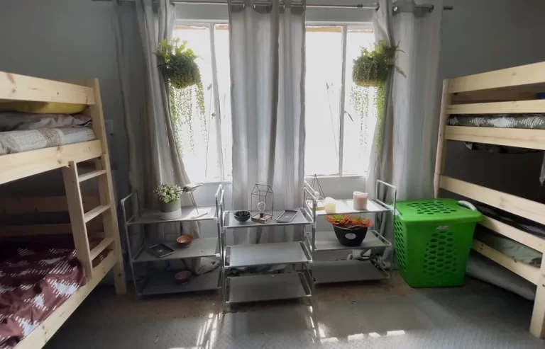 A room with two sets of bunk beds, a window with hanging plants, and a green laundry basket. Between the beds, there are shelving units holding various items. Light filters through curtains.