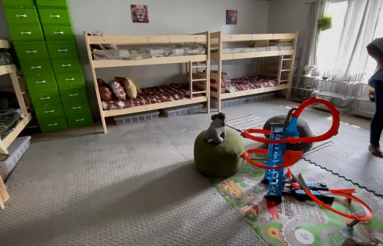 A room with two wooden bunk beds, a green storage cabinet, a toy racetrack set on the floor, and a stuffed animal on a green bean bag.