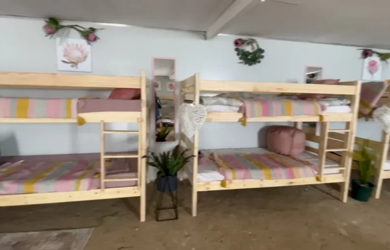 A dorm room with six wooden bunk beds, striped bedding, wall decorations, and plants. The room has a simple, cozy décor with light walls and a cement floor.