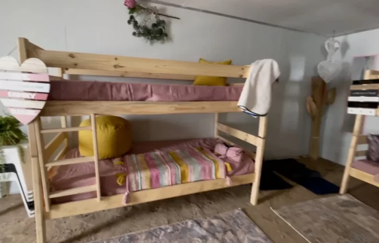 A room with a wooden bunk bed; the top bunk has a pink mattress and a blanket draped over the railing, while the bottom bunk has a striped blanket and a large yellow pillow. Decorative items are on the walls.