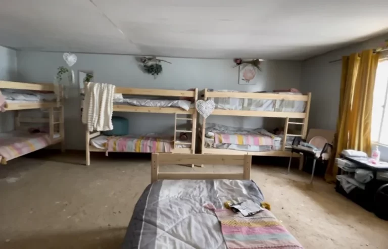 A room with multiple bunk beds, each with different bedding, along with a single bed in the foreground. The room includes various personal items and decorations, with sunlight coming through a window.