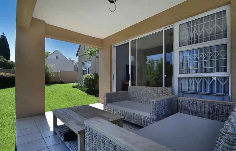 A covered patio area with wicker furniture, including a sofa, chairs, and a coffee table. It opens to a green lawn with a neighboring house in the background.