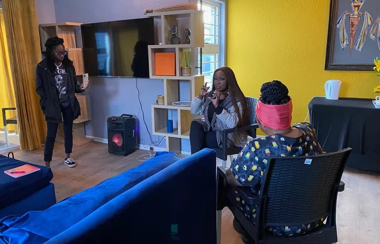 Three people are indoors having a conversation. One person is standing with a cup, while two others are seated with one gesturing. The room has a television, shelves, and a vibrant yellow wall