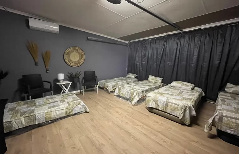 A room with four single beds arranged in a row, each with patterned bedding. There are chairs, a small table with a lamp, and wall decorations. The walls are dark, and the room has wooden flooring.