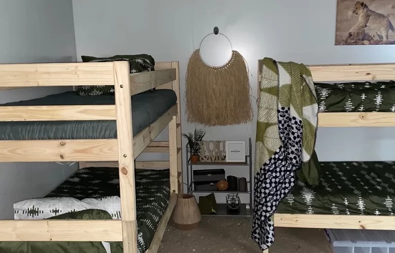 A room with two wooden bunk beds, green and white bedding, a small shelf with decor items, and a circular mirror with a fringed border hanging on the wall. A picture of a lion is displayed above the beds.