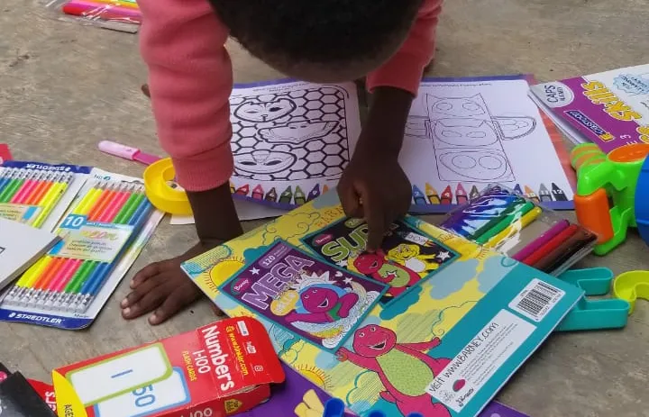 A child is coloring in a coloring book, surrounded by various colored markers, crayons, and additional coloring books on the floor.