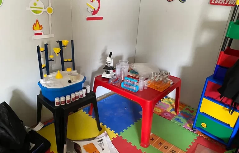 Colorful playroom with a small science lab setup including a microscope, test tubes, and various containers on red and black tables. Walls feature educational decals, and floors have puzzle-piece mats.