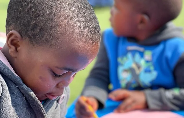 Two young children are seated, focusing intently. One child in the foreground is looking down, holding a pencil. The other child in the background is looking to the side.