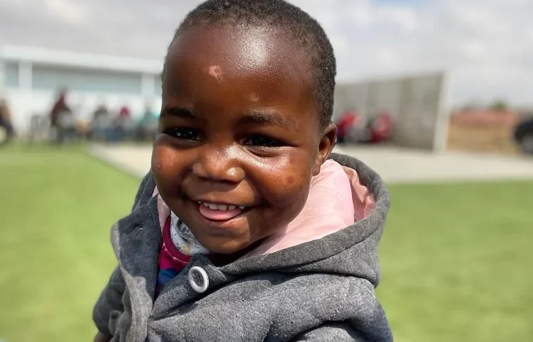 A young child wearing a grey hoodie smiles broadly while standing outdoors on a grassy field with a blurred background of buildings and people.