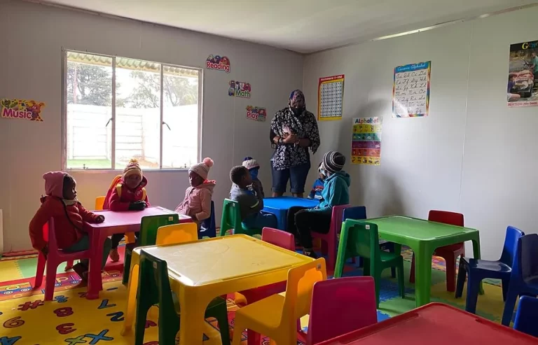 A teacher stands in a classroom with young children sitting at colorful tables. The walls are decorated with educational posters.