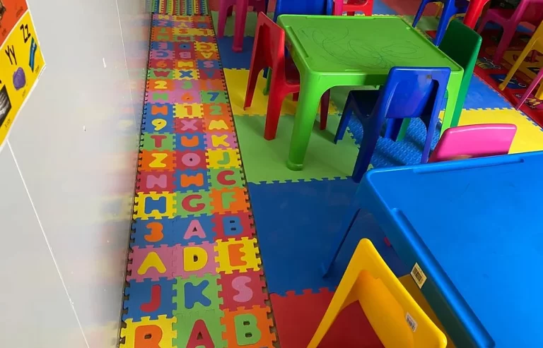 A brightly colored classroom with foam letter and number mats on the floor, and various colored tables and chairs arranged neatly.