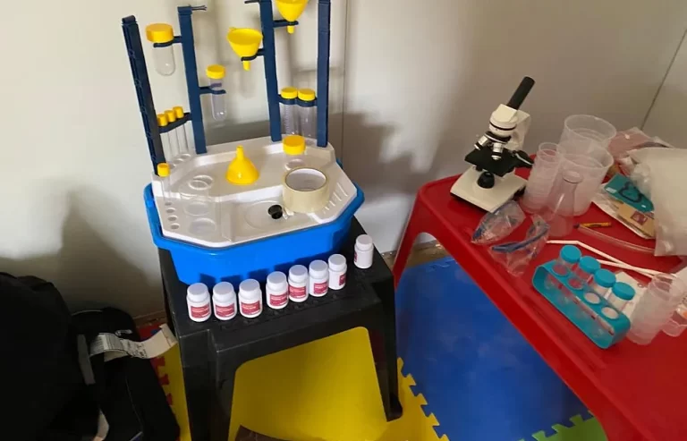 A children's science lab setup with test tubes, a small microscope, plastic bottles, and other lab equipment arranged on a small table and stool. The floor is covered with colorful foam mats.