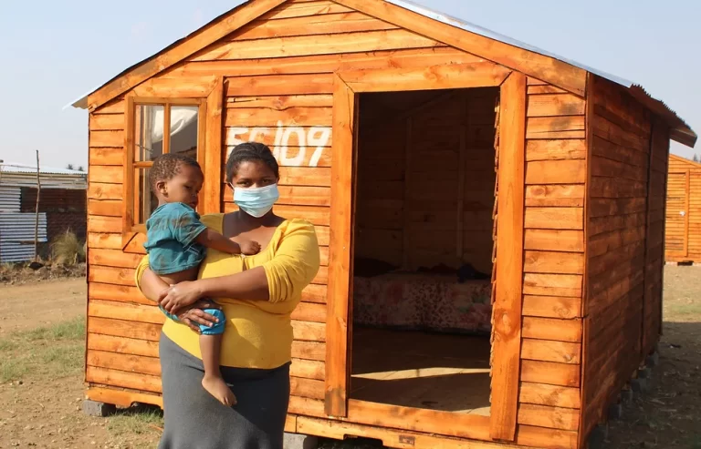 A woman, wearing a mask, stands holding a child in front of a wooden Tiny Home with an open door. The Tiny Home has the number 55109 painted on it.