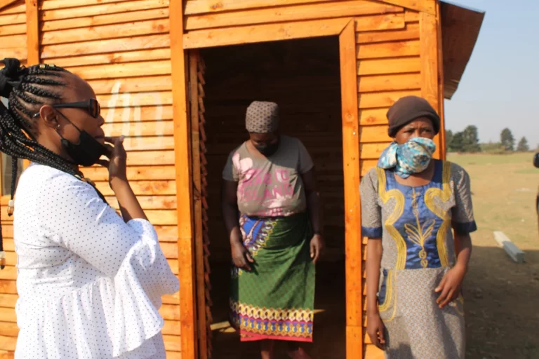 Three individuals standing outside a wooden structure. One person in a white shirt, two others in colorful attire, one wearing a headscarf and another in a face mask.