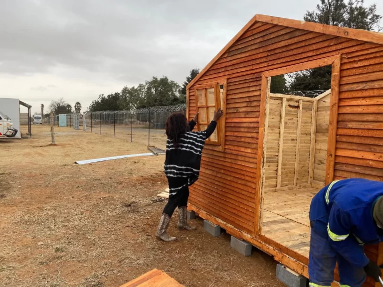 A person is working on installing a window in a small wooden structure while another person, dressed in work attire, is bent over working inside. The structure is set on cinder blocks in an open area.